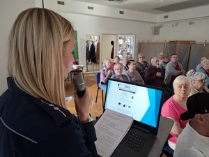 w sali klubu osiedlowego BAZA w Lęborku przy mównicy stoi policjantka, która przemawia przez mikrofon. Przed nią stoi otwarty laptop z prezentacją. Na krzesłach w rzędach siedzą seniorzy