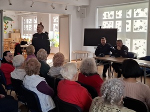 w sali klubu osiedlowego przed siedzącymi na krzesłach w rzędach seniorami stoi policjant. Za nim przy stole siedzą dwaj inni policjanci