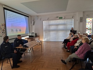 w sali klubu osiedlowego na krzesłach w rzędach siedzą seniorzy. Obok przy mównicy stoi policjantka. Przed seniorami przy stole siedzą dwaj policjanci. Nad ich głowami na ekranie zawieszonym na ścianie wyświetla się prezentacja