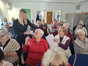 w sali klubu osiedlowego na krzesłach w rzędach siedzą seniorzy. Pomiędzy nimi chodzi policjantka i rozdaje seniorom ulotki
