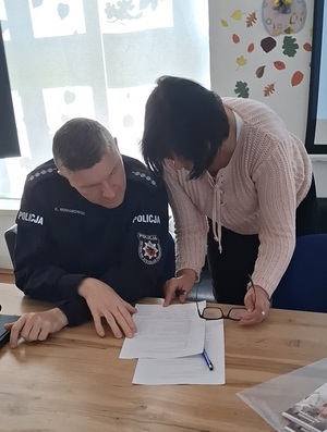 w sali klubu osiedlowego  przy stole pod oknem siedzi policjant. Obok niego stoi kobieta, która nachyla się w jego stronę. Oboje patrzą na kartkę leżącą na stole