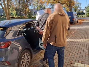mężczyzna z rękoma skutymi za plecami kajdankami, który wyszedł z tylnego siedzenia nieoznakowanego radiowozu. Przed nim stoi nieumundurowany policjant