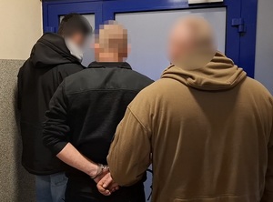 w budynku komendy policji przed drzwiami stoi mężczyzna z rękoma skutymi za plecami kajdankami. Przy nim stoi dwóch nieumundurowanych policjantów