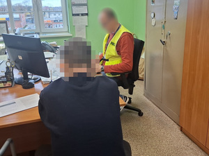przy biurku przed komputerem siedzi policjant, który przesłuchuje siedzącego obok na krześle zatrzymanego mężczyznę