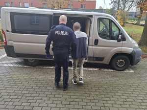 przed otwartymi bocznymi drzwiami nieoznakowanego radiowozu typu bus stoi policjant, który trzyma za ramię zatrzymanego mężczyznę