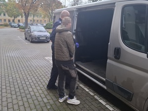 przed otwartymi bocznymi drzwiami nieoznakowanego radiowozu służby konwojowej stoi mężczyzna, który ma ręce skute z przodu kajdankami. Przy nim stoi policjant