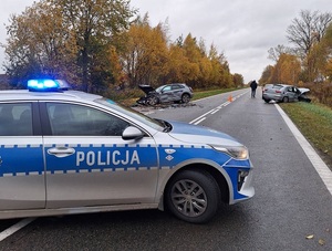 miejsce wypadku drogowego. Po obu stronach jezdni na poboczu znajdują się dwa osobowe samochody: audi i seat. Oba są uszkodzone, mają rozbity przód i boki. Na pierwszym planie, w poprzek jezdni jest ustawiony policyjny oznakowany radiowóz