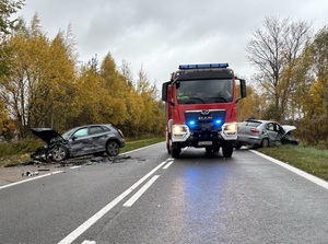 miejsce wypadku drogowego. Po obu stronach jezdni na poboczu znajdują się dwa osobowe samochody: audi i seat. Oba są uszkodzone, mają rozbity przód i boki. Na środku jezdni, pomiędzy samochodami znajduje się wóz straży pożarnej