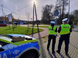 w okolicy cmentarza, przed oznakowanym radiowozem stoją dwie policjantki ruchu drogowego. Chodnikiem idą przechodnie, drogą dla rowerów jedzie rowerzysta