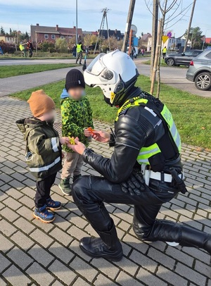 w okolicy cmentarza policjant ruchu drogowego ubrany w kombinezon motocyklowy, z kaskiem na głowie przekazuje dwóm małym chłopcom breloczki odblaskowe