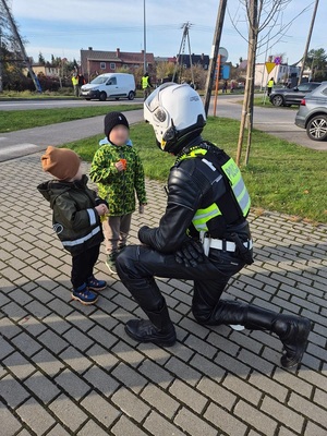 w okolicy cmentarza policjant ruchu drogowego ubrany w kombinezon motocyklowy, z kaskiem na głowie klęczy na jednym kolanie przed dwoma małymi chłopcami, trzymającymi po breloczku odblaskowym