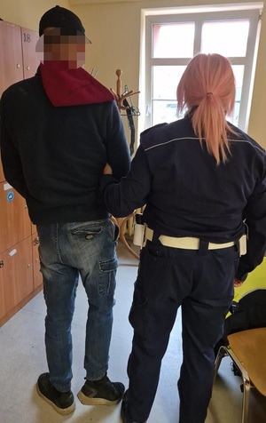 w pomieszczeniu stoi policjantka ruchu drogowego, która trzyma za ramię zatrzymanego mężczyznę. Oboje są zwróceni tyłem do obiektywu