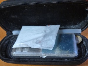 otwarte czarne etui. W środku znajduje się mała waga elektroniczna, na której leży woreczek strunowy wypełniony białym proszkiem oraz plastikowa rurka