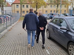 nieumundurowany policjant prowadzi na zewnątrz budynku komendy mężczyznę, który ma ręce skute za plecami kajdankami