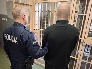 w okratowanym pomieszczeniu przed okratowanymi drzwiami stoi policjant służby konwojowej, który trzyma za ramię zatrzymanego mężczyznę