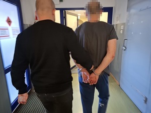 nieumundurowany policjant prowadzi korytarzem mężczyznę, który ma ręce skute za plecami kajdankami