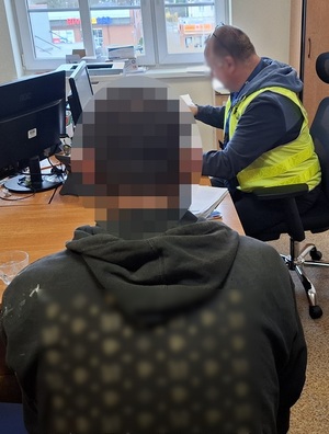 w pokoju przesłuchań na krześle siedzi zatrzymany mężczyzna, zwrócony tyłem do obiektywu. Przed nim przy biurku przed komputerem siedzi policjant