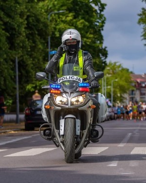 oznakowanym motocyklem jedzie policjant. Za nim widać biegnących uczestników zawodów sportowych