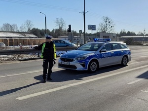 na pasie ruchu stoi policjantka ruchu drogowego, która trzyma wyciągniętą przed siebie tarczę do zatrzymywania pojazdów. Za nią przy skrzyżowaniu stoi oznakowany radiowóz