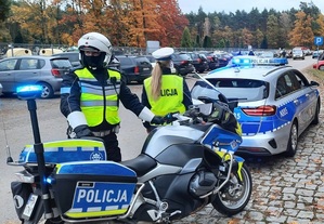 przed cmentarzem, na miejscu parkingowym znajduje się dwoje policjantów ruchu drogowego: policjant przy oznakowanym policyjnym motocyklu i odwrócona do niego tyłem policjantka stojąca przy oznakowanym radiowozie