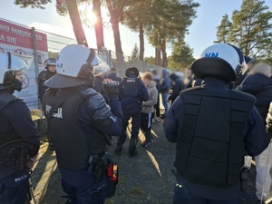 przed wejściem na stadion policjanci ubrani w sprzęt przeciwuderzeniowy otaczają kibiców. Jeden z policjantów trzyma kamerę