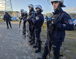 Przed stadionem stoją policjanci ubrani w sprzęt przeciwuderzeniowy. Policjanci trzymają strzelby gładkolufowe