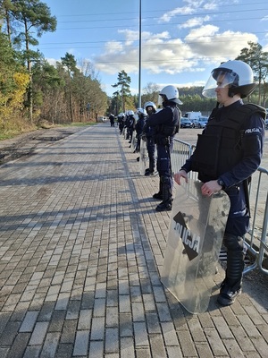 z boku stadionu stoją w rzędzie policjanci ubrani w sprzęt przeciwuderzeniowy. Trzymają tarcze
