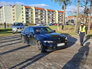 samochody osobowe, wjeżdżające jeden za drugim na nieutwardzoną drogę obok stadionu od strony lasu. Na drodze stoi policjantka ruchu drogowego obserwująca pojazdy. W tle, na ulicy stoją dwaj policjanci ruchu drogowego, którzy kierują pojazdy na tę drogę