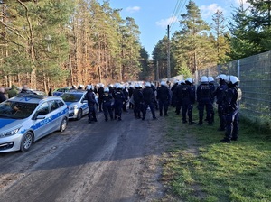 przy płocie okalającym stadion, od strony kibiców przyjezdnych, stoi kilkudziesięciu policjantów ubranych w sprzęt przeciwuderzeniowy. Otaczają kibiców. Wzdłuż drogi stoją policyjne radiowozy