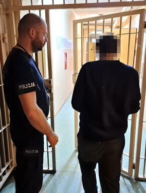 zatrzymany mężczyzna przechodzi przez okratowane drzwi pomieszczenia dla osób zatrzymanych. Obok stoi policjant służby dyżurnej