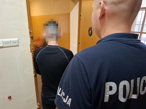 w celi stoi zatrzymany mężczyzna. Za nim przy otwartych drzwiach celi stoi policjant służby dyżurnej