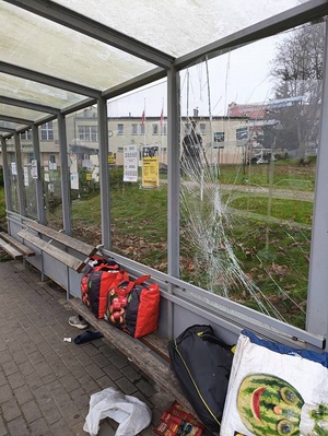 wiata przystanku autobusowego z pękniętą szybą. Na ławeczce leżą torby z bagażem, pod nią leży but i klapek