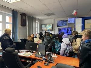 w pomieszczeniu dyżurnego policji stoi grupa uczniów przyglądającym się zawieszonym na ścianie monitorom. Za nimi stoi policjantka
