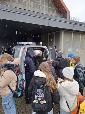 za radiowozem stoi grupa uczniów. Z tylnej części radiowozu wychodzi jeden z uczniów. Obok stoi policjantka