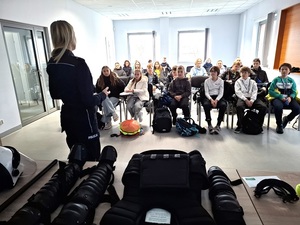 w auli lęborskiej komendy policji siedzą na krzesłach uczniowie. Przed nimi, przy stole, na którym leży policyjny sprzęt przeciwuderzeniowy, stoi policjantka