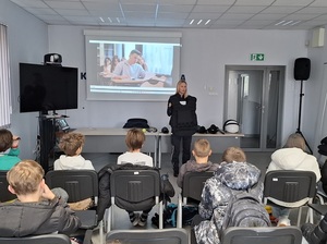 w auli lęborskiej komendy policji siedzą na krzesłach uczniowie. Przed nimi, przy stole, na którym leży policyjny sprzęt przeciwuderzeniowy, stoi policjantka. Policjantka trzyma w rękach kamizelkę przeciwuderzeniową. Za nią, na ekranie wyświetla się film profilaktyczny