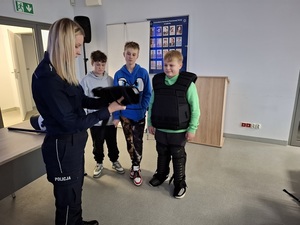 w auli lęborskiej komendy policji przed policjantką stoi trzech uczniów. Jeden jest częściowo ubrany w sprzęt przeciwuderzeniowy. Policjantka trzyma w rękach nałokietnik