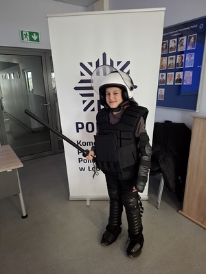 w auli lęborskiej komendy policji na tle roll up z napisem Komenda Powiatowa Policji w Lęborku stoi chłopiec ubrany w sprzęt przeciwuderzeniowy