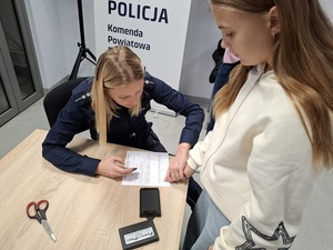 w auli lęborskiej komendy policji przy stole siedzi policjantka. Przed nią stoi uczennica. Policjantka przykłada jej palec do karty daktyloskopijnej