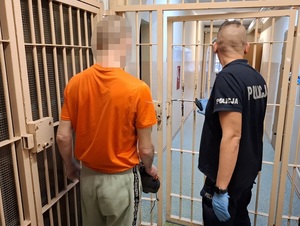 w korytarzu pomieszczenia dla osób zatrzymanych przed otwartymi okratowanymi drzwiami stoi zatrzymany mężczyzna. Obok stoi policjant służby dyżurnej