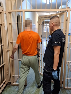 w korytarzu pomieszczenia dla osób zatrzymanych przez otwarte okratowane drzwi przechodzi zatrzymany mężczyzna. Obok stoi policjant służby dyżurnej