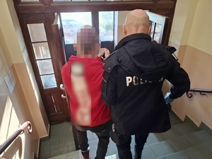 policjant służby konwojowej prowadzi schodami w dół w kierunku wyjścia z budynku komendy mężczyznę, który ma założone kajdanki zespolone