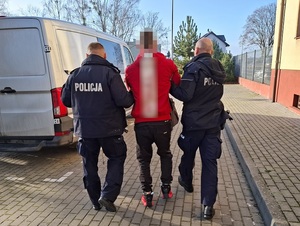 dwa policjanci służby konwojowej prowadzą w stronę nieoznakowanego radiowozu mężczyznę, który ma założone kajdanki zespolone