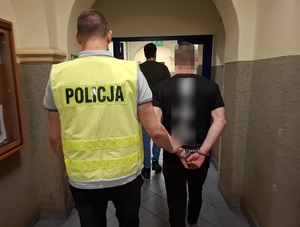 policjant w kamizelce odblaskowej z napisem policja prowadzi korytarzem mężczyznę, który ma ręce skute za plecami kajdankami