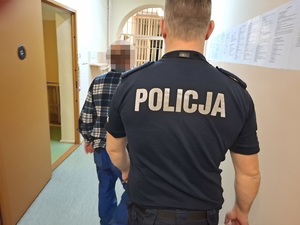 korytarzem pomieszczenia dla osób zatrzymanych idzie mężczyzna. Za nim idzie policjant służby dyżurnej