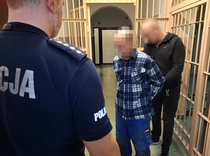 w korytarzu pomieszczenia dla osób zatrzymanych nieumundurowany policjant zakłada kajdanki na ręce mężczyzny. Przed mężczyzną stoi policjant służby dyżurnej
