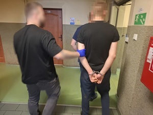 przed otwartymi drzwiami prowadzącymi na korytarz pomieszczenia służbowego stoi mężczyzna, który ma ręce skute za plecami kajdankami. Za przedramię trzyma go policjant kryminalny