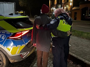 pora wieczorowa. Za radiowozem stoją obok siebie policjantka ruchu drogowego i starszy mężczyzna. Policjantka obejmuje ramieniem mężczyznę
