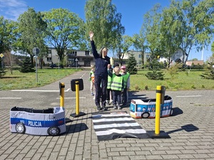 przed zabawkowym przejściem dla pieszych stoi policjantka z grupą przedszkolaków. Policjantka podnosi rękę w górę