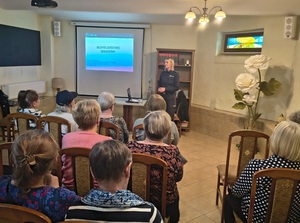 w sali na krzesłach siedzą seniorzy. Przed nimi na tle wyświetlanej na ścianie prezentacji, stoi policjantka